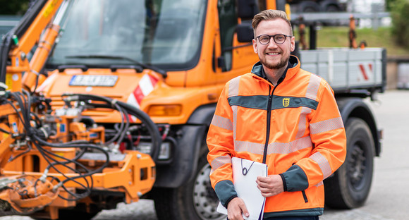 Ein Mann steht in organgfabener Arbeiterjacke vor einem Mähfahrzeug der Straßenmeisterei.